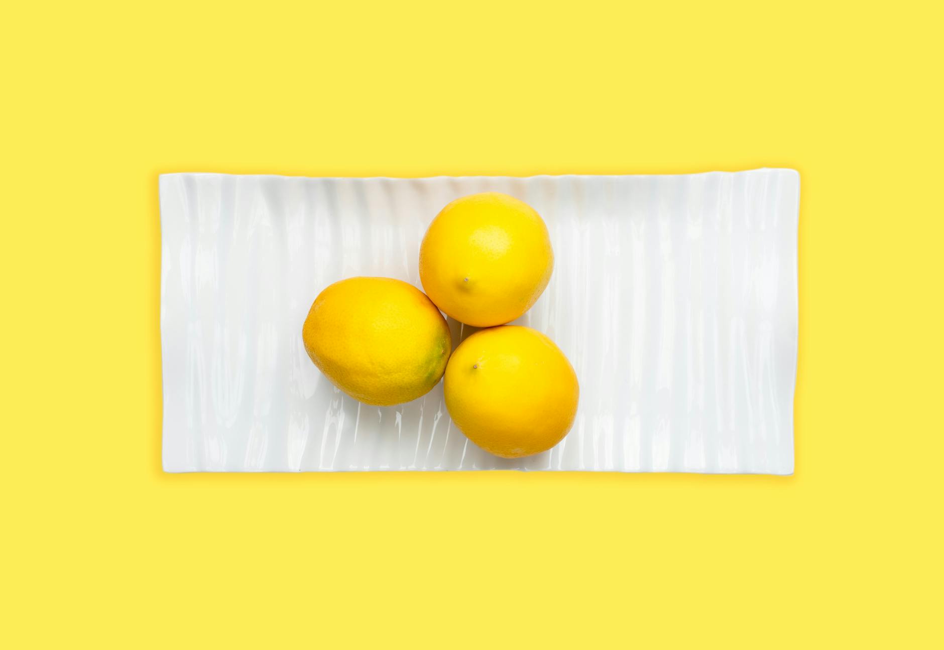 A hand with white nails holding a lemon on a soft pink background, surrounded by three additional lemons
