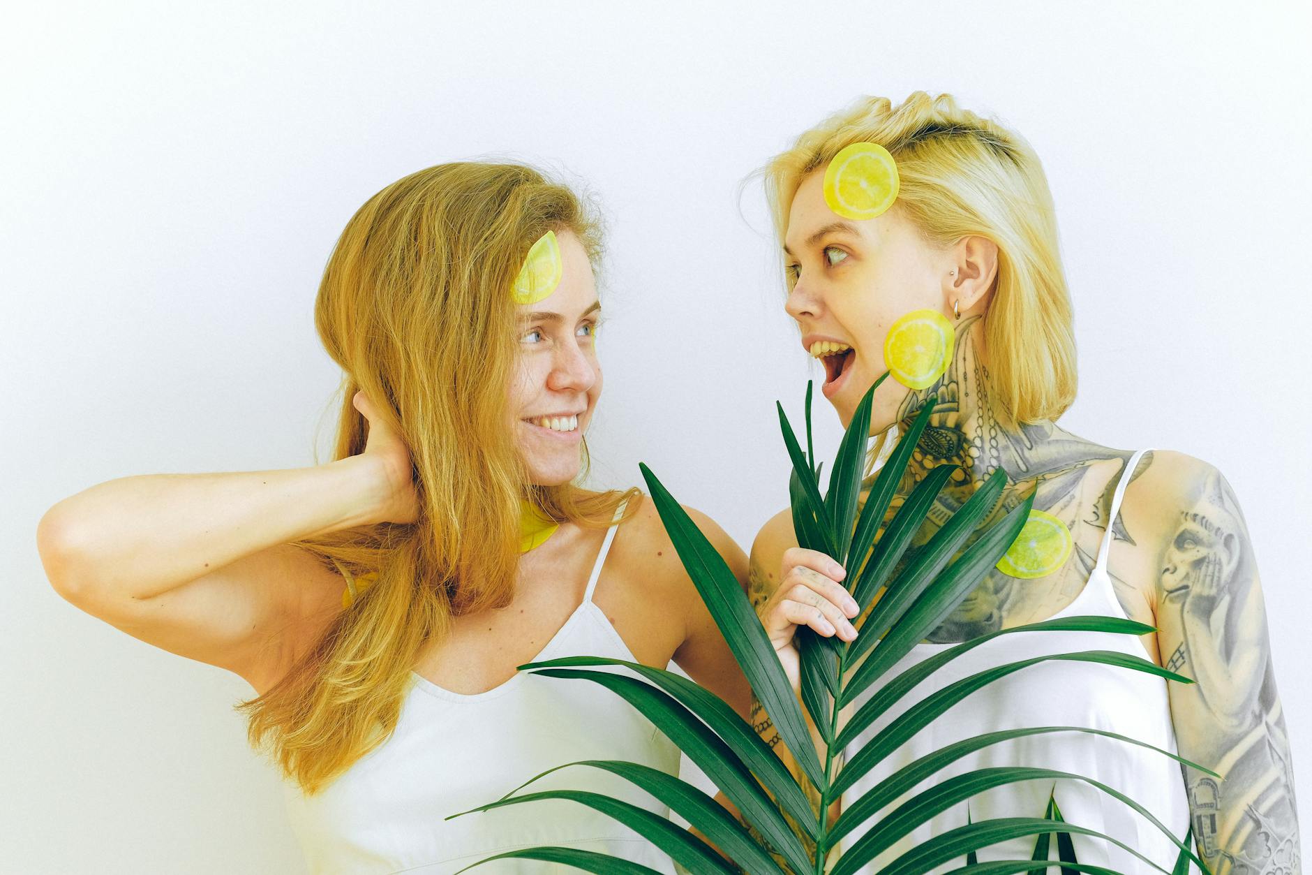 Two smiling women with lemon slices and tropical plants, expressing joy and connection indoors.