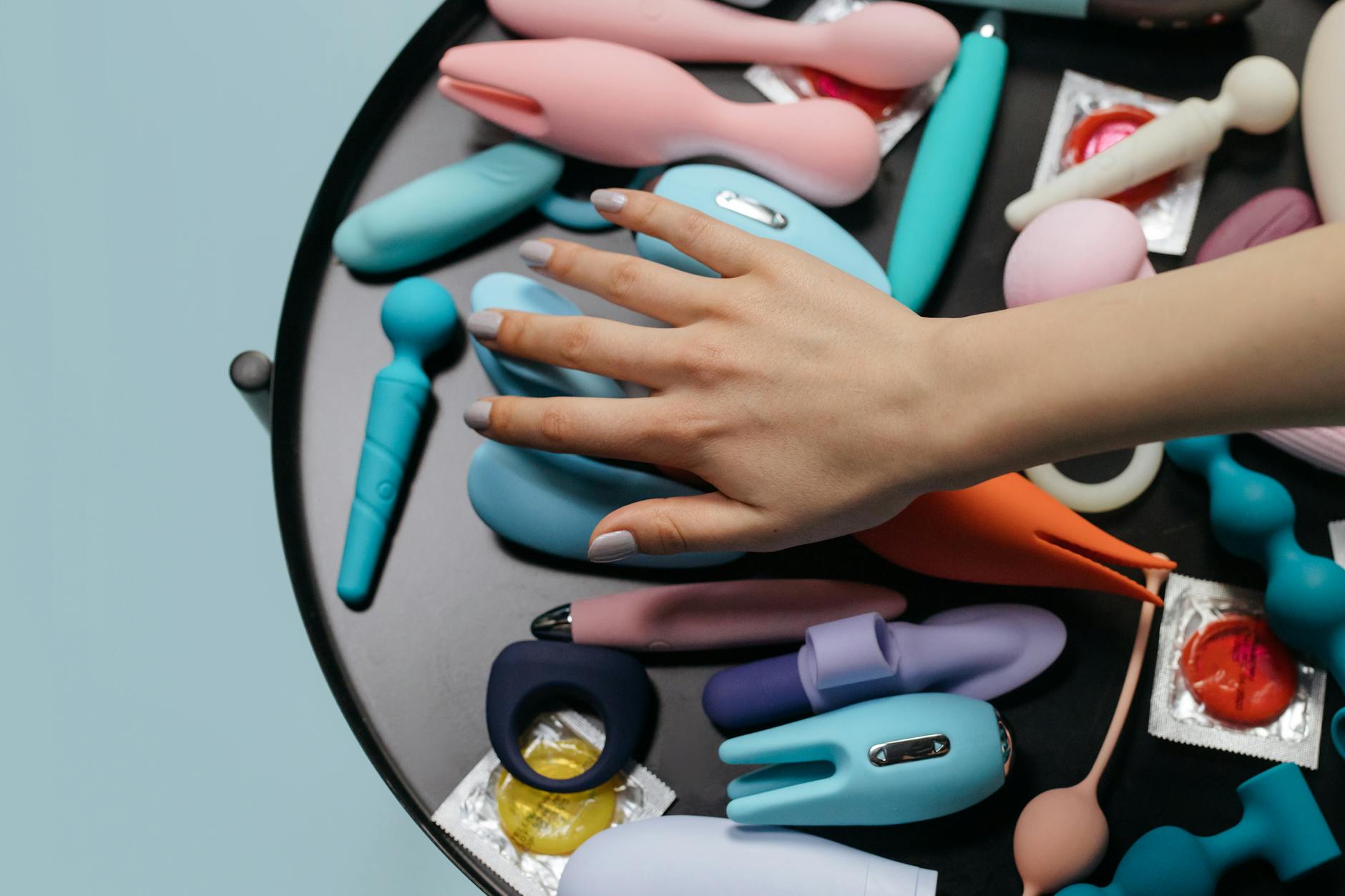 Two hands holding pink and blue silicone clitoral vibrators against a pastel background
